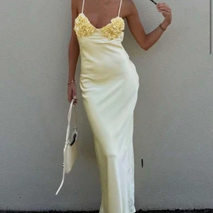 Ljusgul långklänning med volangdetaljer - Säljer en elegant ljusgul långklänning i satin med smala axelband och vackra volangdetaljer över bysten. Klänningen har öppen rygg med knyt och en figurnära passform som ger en stilren siluett. Perfekt för sommarens speciella tillfällen!