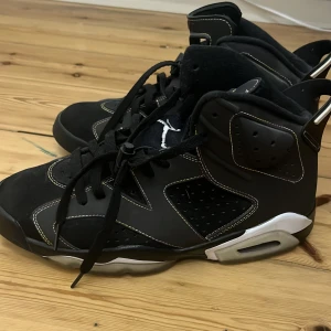 Svarta Nike Air Jordans 6 Retro - Säljer ett par svarta Nike Air Jordans 6 Retro med vita och grå detaljer. Skorna har klassisk hög siluett, perforerade paneler och snörning framtill. Ikonisk Jumpman-logga syns på hälen och tungan. Perfekta för dig som gillar streetwear och sneakerskultur.