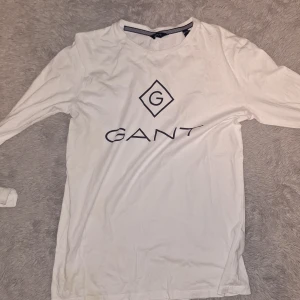 Vit långärmad tröja från GANT - Säljer en vit långärmad tröja från GANT med stor logga och tryck framtill. Tröjan har rund halsringning och är i ett mjukt bomullsmaterial. Perfekt för en stilren och enkel look.