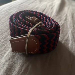 Flätat bälte med läderdetaljer - Säljer ett snyggt flätat bälte i mörkblått och rött med bruna läderdetaljer och silverspänne. Perfekt accessoar för att lyfta din outfit med lite färg och stil.