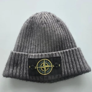 Stone Island mössa grå - Snygg grå ribbstickad mössa från Stone Island med uppvikt kant och klassisk logotyp framtill. Perfekt för alla dagar och enkel att matcha med olika outfits. Rep