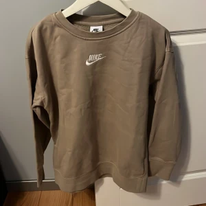 Beige sweatshirt från Nike - Säljer en beige sweatshirt från Nike med vit broderad logga på bröstet. Tröjan har rund halsringning och långa ärmar. Perfekt för en avslappnad och sportig stil.