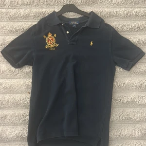 Marinblå pikétröja från Polo Ralph Lauren - Säljer en klassisk marinblå pikétröja från Polo Ralph Lauren med broderad gul logga på bröstet och en stor broderad detalj med siffran 3. Tröjan har krage och korta ärmar, perfekt för en stilren look.