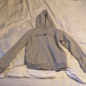Grå hoodie från Sail Racing - Säljer en ljusgrå hoodie från Sail Racing med svart logga på bröstet. Tröjan har huva, långa ärmar och ribbade muddar vid ärmslut och nederkant. Perfekt för en avslappnad stil. Priset kan diskuteras 
