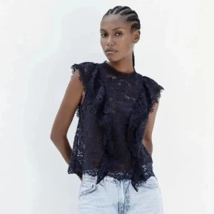 Svart spetsblus med volang - Säljer en svart ärmlös blus i spets med volangdetaljer framtill. Blusen har rund halsringning och knäppning bak i nacken. Perfekt att styla med jeans eller kjol för en trendig look.