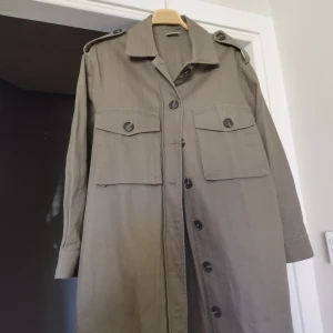 Olivgrön lång rock med axelklaffar - Säljer en olivgrön lång rock med klassisk krage och stora bröstfickor med knappar. Jackan har axelklaffar och stängs med knappar framtill. Perfekt för dig som gillar en stilren och tidlös look.