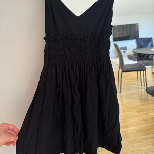Svart kortklänning med v-ringning - Säljer en svart kortklänning med smala axelband och v-ringad hals. Klänningen har smockad midja som ger en snygg passform och ett lätt, fladdrigt tyg. Perfekt för varma dagar eller att styla med en jacka.