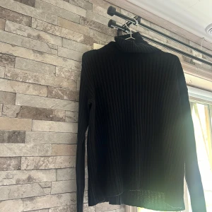 Svart ribbad polotröja från Hennes Collection - Säljer en svart ribbad polotröja från Hennes Collection. Tröjan har lång ärm och är tillverkad i ett stretchigt, stickat material som sitter snyggt på kroppen. Perfekt för lager-på-lager eller som den är.