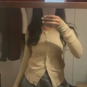 Beige stickad kofta  - Säljer en beige kofta med stora dekorativa knappar framtill. Tröjan har lång ärm och är figurnära i modellen. Perfekt att styla till både jeans och kjol.