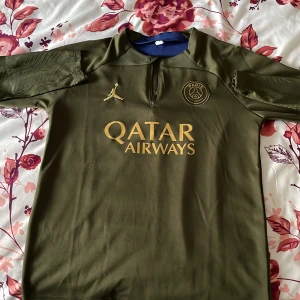 PSG fotbollströja i olivgrönt med guldtryck - Säljer en olivgrön Paris Saint-Germain fotbollströja med guldiga detaljer och Qatar Airways-tryck på bröstet. Tröjan har korta ärmar, dragkedja vid halsen och PSG-logga samt Jordan-logga framtill.
