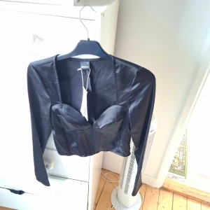Svart långärmad korsettopp från Gina Tricot - Säljer en svart långärmad korsettopp från Gina Tricot. Toppen har markerade kupor, croppad passform och är tillverkad i ett glansigt material som ger en lyxig känsla. Perfekt för att lyfta din outfit!