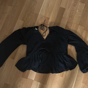 Svart blus med knyt från H&M - Säljer en svart blus från H&M med v-ringning och knyt i nacken. Blusen har långa ärmar och volang nertill vilket ger en snygg siluett. Perfekt för dig som gillar stilrena plagg med fina detaljer.