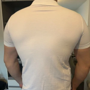 Vit pikétröja från Polo Ralph Lauren - Säljer en klassisk vit pikétröja från Polo Ralph Lauren med röd broderad logga på bröstet. Tröjan har korta ärmar, krage och knappar vid halsen. Storlek L