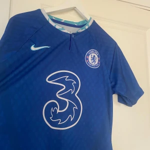 Chelsea FC hemmatröja 2022/23 - Säljer en blå Chelsea FC fotbollströja från Nike med klubbmärke och sponsorlogga på bröstet. Tröjan har korta ärmar och ett mönstrat innerkrage i ljusblått och vitt. Perfekt för fans som vill visa sitt stöd på matchdag!