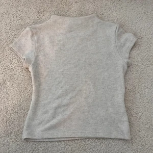 Beige kortärmad topp från Pull&Bear - Säljer en ljusgrå, tight och mjuk topp från Pull&Bear. Toppen har korta ärmar och en något högre halsringning. Perfekt att matcha med jeans eller kjol för en enkel och stilren look.