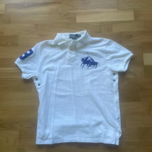 Ralph lauren piké - Vit pikétröja från Polo Ralph Lauren med klassisk krage och knappar. Stor blå logga på bröstet och siffran 3 på ärmen. Perfekt för en sportig och stilren look.