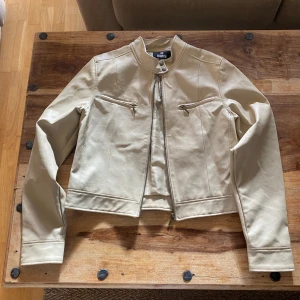 Beige skinnjacka från Cam - Säljer en beige skinnjacka från Cam med ståkrage och dragkedja framtill. Jackan har två snedställda fickor med dragkedja och detaljerade sömmar. Perfekt för dig som gillar en stilren och enkel look.