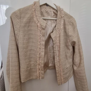 Beige tweed kavaj med fransdetaljer - Säljer en beige kavaj i tweed med fransdetaljer längs kanterna. Jackan har lång ärm, rak passform och är fodrad. Perfekt för en dressad look och passar till många olika outfits. 
