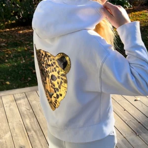 Vit hoodie med leopardmönstrad paljettdekor - Vit hoodie med huva och en stor dekorativ leopard i guld- och svartpaljetter på ryggen. Tröjan har en avslappnad passform och är perfekt för dig som vill sticka ut med ett coolt djurmotiv.