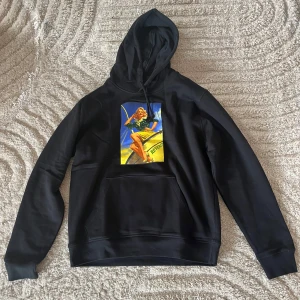 Svart hoodie med retrotryck - Svart hoodie med stor huva och magficka. På bröstet finns ett färgglatt retrotryck med en pinup-tjej i grönt och gult. Tröjan har en klassisk passform och är perfekt för en avslappnad stil.