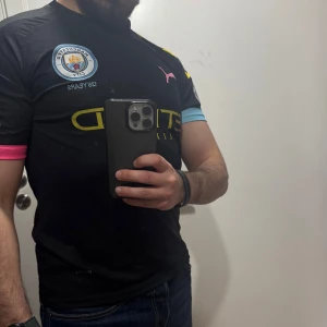 Manchester City fotbollströja Mahrez 26 - Säljer en svart Manchester City fotbollströja med Mahrez och nummer 26 på ryggen. Tröjan har klubbmärke på bröstet, Puma-logga och färgdetaljer i blått och rosa på ärmsluten. Perfekt för fans av Premier League och Mahrez.