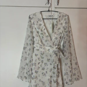 Blommig omlottklänning från NA-KD - Superfin vit omlottklänning från NA-KD med blommigt mönster i lila, grönt och rosa. Klänningen har långa ärmar, v-ringning och knyt i midjan. Perfekt för vår och sommar, lätt och luftig i polyester. Storlek 38. Aldrig använd med prislapp kvar