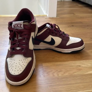 Nike Dunk Low Bordeaux/Vit - Nike Dunk Low sneakers i bordeauxrött och vitt. Storlek 38, har växt ut skorna därför säljer jag dom. Skorna har en svart repa på sulan.