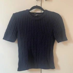 Kabelstickad halvärmad tröja från Gina Tricot - Säljer en mörkblå stickad tröja från Gina Tricot med korta ärmar och klassiskt kabelstickat mönster. Tröjan har rund halsringning och sitter snyggt med en normal passform. Perfekt till jeans eller kjol. 