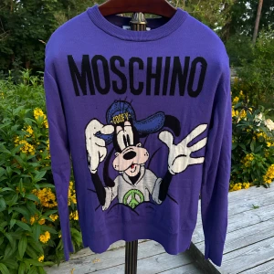 Lila Moschino x H&M Goofy tröja XS - Supercool lila stickad tröja från Moschino x H&M i samarbete, med stor Goofy-illustration fram och bak. Tröjan har rund hals, långa ärmar och en relaxed vibe. Perfekt statement piece för dig som gillar färg och Disney. Dem ända defekterna ser ni på bilderna.