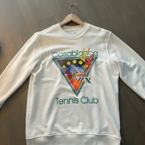 Vit sweatshirt Casablanca Tennis Club - Säljer en vit sweatshirt från Casablanca med färgglatt tryck framtill och texten 'Tennis Club'. Tröjan har rund halsringning och långa ärmar. Perfekt för dig som gillar unika prints och sportig stil.