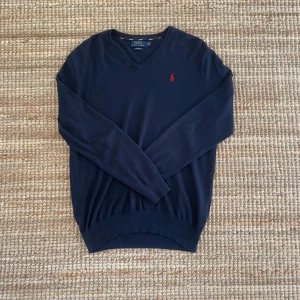 Marinblå tröja från Polo Ralph Lauren - Snygg marinblå tröja från Polo Ralph Lauren i mjuk pima bomull. Klassisk v-ringning och långärmad modell med den ikoniska röda loggan broderad på bröstet. Perfekt för dig som gillar stilrena och tidlösa plagg.