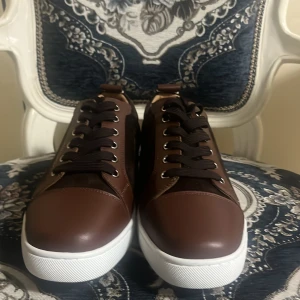 Bruna sneakers från Louboutin - Snygga bruna sneakers från Louboutin med klassisk röd sula och vita kanter. Skorna har ovandel i skinn och mocka, svarta snören och rund tå. Perfekta för dig som vill sticka ut med exklusiv stil och detaljer. Helt nya aldrig användt, storlek 42 men ja h ar storlek 43 och dem var lite stora så kommer passa 43,5 Eller 44. Pris kan diskuteras. Dem kommer med låda och dust bag.