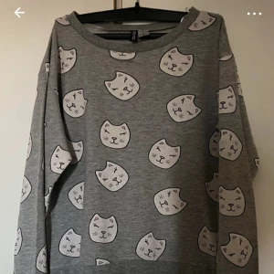 Grå sweatshirt med kattmönster H&M XS - Grå sweatshirt från H&M i storlek XS med gulligt mönster av vita kattansikten över hela tröjan. Mjuk bomullskänsla, rund hals och långa ärmar. Perfekt för dig som gillar söta prints och vill ha en chill vibe.