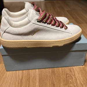 Vita sneakers från Lanvin med färgglada snören - Snygga vita sneakers från Lanvin med coola, breda snören i rött, svart, gult och vitt zigzag-mönster. Skorna har en beige sula och är tillverkade i mocka och skinn. Perfekta för dig som vill sticka ut med en exklusiv och trendig look.