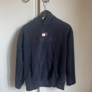 Mörkblå hoodie från Tommy Hilfiger - Snygg mörkblå hoodie från Tommy Hilfiger med klassisk logga broderad på bröstet. Tröjan har huva, känguruficka och långa ärmar. Perfekt för en chill och stilren look. Materialet är mjuk bomull som känns skönt mot huden. Aldrig använt hoodien pris kan diskuteras 
