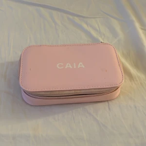 Rosa necessär från CAIA - Söt och praktisk rosa necessär från CAIA med vitt tryck på locket. Väskan har en rektangulär form, dragkedja runt om och är tillverkad i ett slätt syntetmaterial. Perfekt för smink eller småprylar när du är på språng.