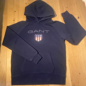 Marinblå hoodie från GANT - Snygg marinblå hoodie från GANT med broderad logga och sköld på bröstet. Klassisk känguruficka framtill och huva. Perfekt för dig som gillar stilren och sportig look. Mjuk bomullskänsla och långa ärmar.