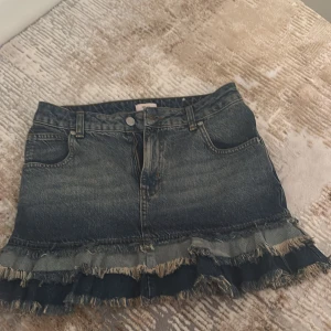 Jeanskjol med volang från Nelly - Mörkblå jeanskjol från Nelly med volang och fransad nederkant. Storlek 32.