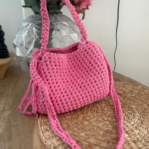Handgjord rosa stickad axelväska - Superfin rosa axelväska som är handgjord och stickad i tjockt garn. Väskan har en flätad axelrem och coola snörningsdetaljer på sidorna. Perfekt för dig som vill ha en unik accessoar med färgklick till din outfit.