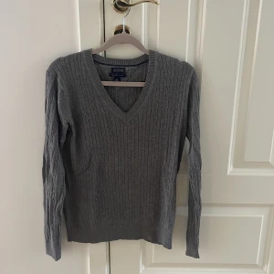 Grå kabelstickad tröja Tommy Hilfiger - Kabelstickad grå tröja från Tommy Hilfiger i 100% pima bomull. V-ringad modell med lång ärm och diskret logga på ärmen. Perfekt för dig som gillar klassisk och stilren look.