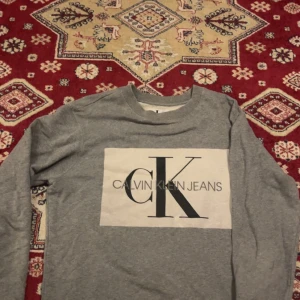 Grå Calvin Klein Jeans sweatshirt - Snygg grå sweatshirt från Calvin Klein Jeans med stort CK-logga framtill. Tröjan har rund hals, långa ärmar och är tillverkad i mjuk bomullsmix. Perfekt för en clean och stilren look.