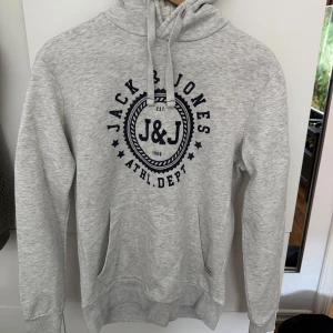 Grå hoodie från Jack & Jones - Snygg ljusgrå hoodie från Jack & Jones med stort J&J-tryck i mörkblått framtill. 