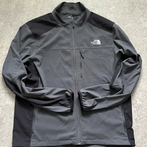Svart och grå vindjacka från The North Face - Snygg svart och grå vindjacka från The North Face med dragkedja framtill och bröstficka med zip. Jackan har hög krage, långa ärmar och logga på både fram- och baksida. Tillverkad i polyester, perfekt för blåsiga dagar.