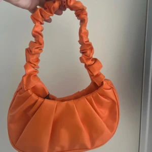  axelväska -  orange axelväska med snygg rynkad rem och rundad form. Väskan har ett slätt, glansigt material och är perfekt för att lyfta vilken outfit som helst. Enkel att bära över axeln och rymmer det viktigaste du behöver ha med dig. Helt ny