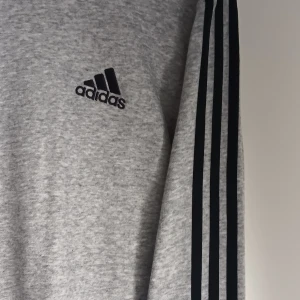Grå långärmad Adidas tröja - Grå långärmad tröja från Adidas med klassiska svarta ränder längs ärmarna och broderad logga på bröstet. Skön passform och tillverkad i mjuk bomullsmix, perfekt för en sportig och avslappnad stil.