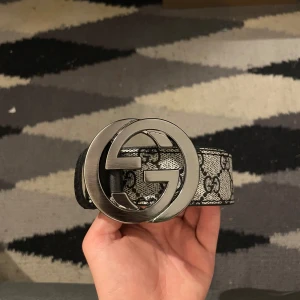 GG bälte med silverfärgat spänne - Snyggt bälte med klassiskt GG-mönster i svart och grått. Bältet har ett stort, rundat spänne i silverfärgad metall med dubbla G-loggan. Remmen är i skinn och mocka, med detaljer i svart läder. Perfekt för att lyfta vilken outfit som helst.