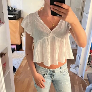 TRENDIG Vit blus med volang och v-ringning - Supersöt vit blus med v-ringning och volangkant. Den har korta, luftiga ärmar och är croppad med en flowy passform. Tunt, lätt bomullstyg som känns perfekt för varma dagar. Snygg spetsdetalj längs halsringningen för extra charm.