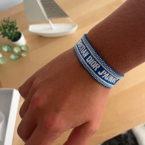 Blått Christian Dior J'Adior armband - Snyggt blått armband från Christian Dior med texten 'J'Adior' broderad i vitt. Armbandet har vita detaljer och avslutas med blå tofsar. Perfekt accessoar för att lyfta din outfit med en lyxig touch och cool streetkänsla.