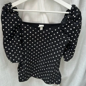 Svart prickig topp med puffärm H&M - Svart topp från H&M med vita prickar och trendiga puffärmar. V-ringad halsringning och figurnära passform. Perfekt för dig som gillar en lekfull och stilren look. Toppens material är mjukt och stretchigt för extra komfort.