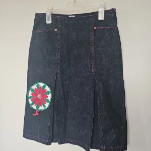Jeanskjol med broderad detalj - Mörk jeanskjol med röda kontrastsömmar och två bakfickor. På framsidan finns en stor, broderad blomma i rött, grönt och vitt som ger kjolen en unik vibe. Kjolen har dragkedja i sidan och lätt utställd form som slutar strax ovanför knäet.